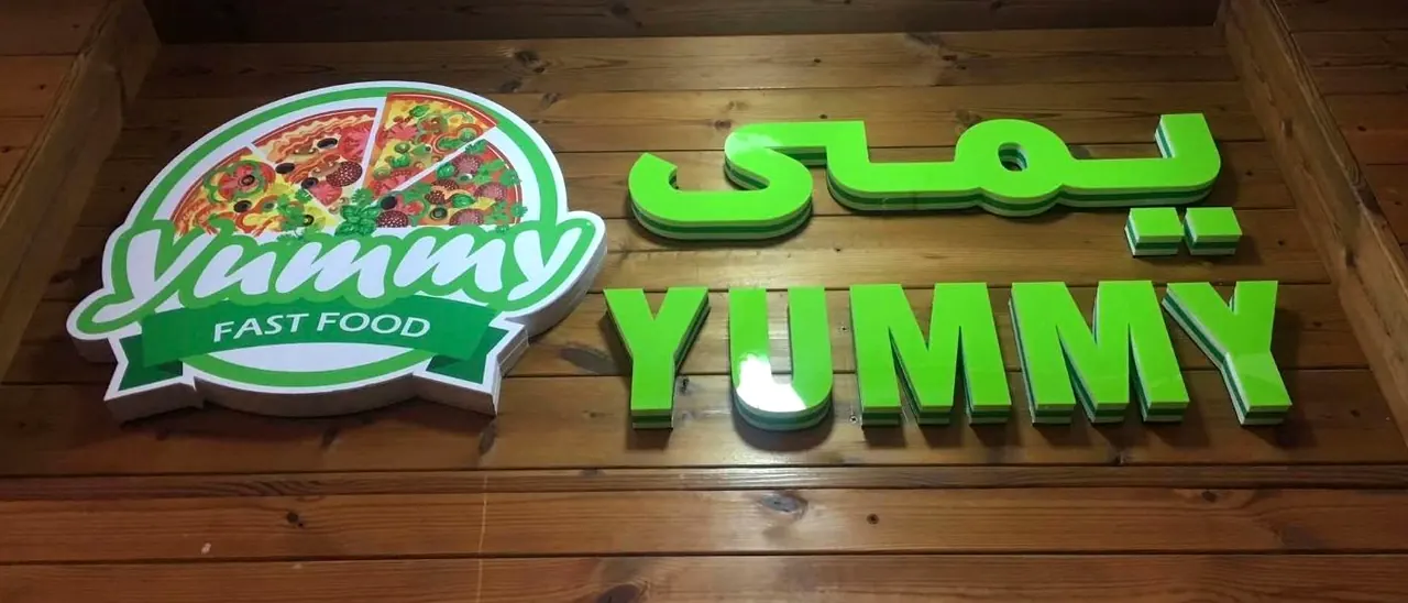 Bright green layered acrylic 3D letters spelling Yummy in English and Dari beside a printed pizza logo, on a timber wall.